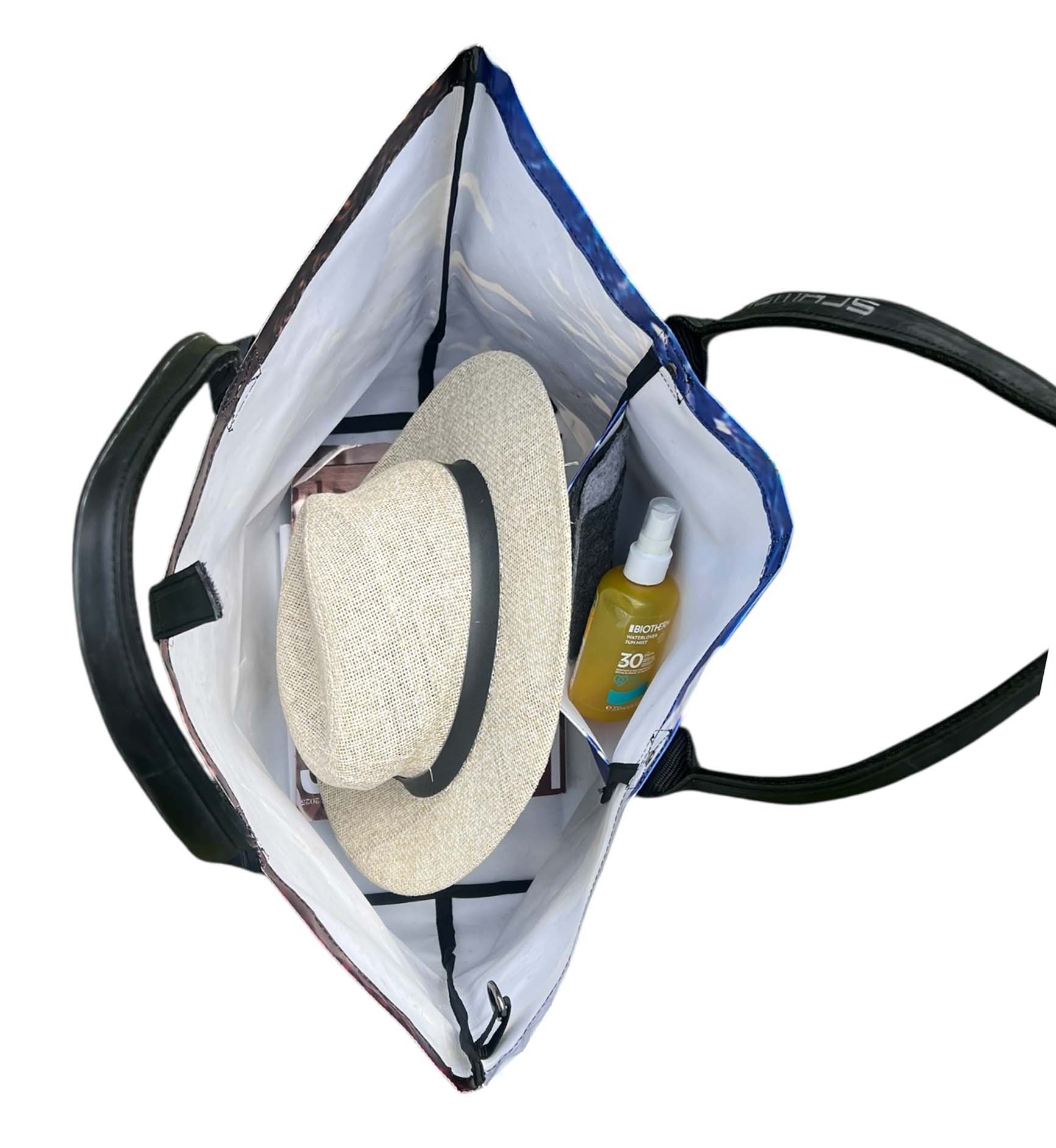 Open tote bag with a sun hat, sunglasses, and a bottle of sunscreen on a white background