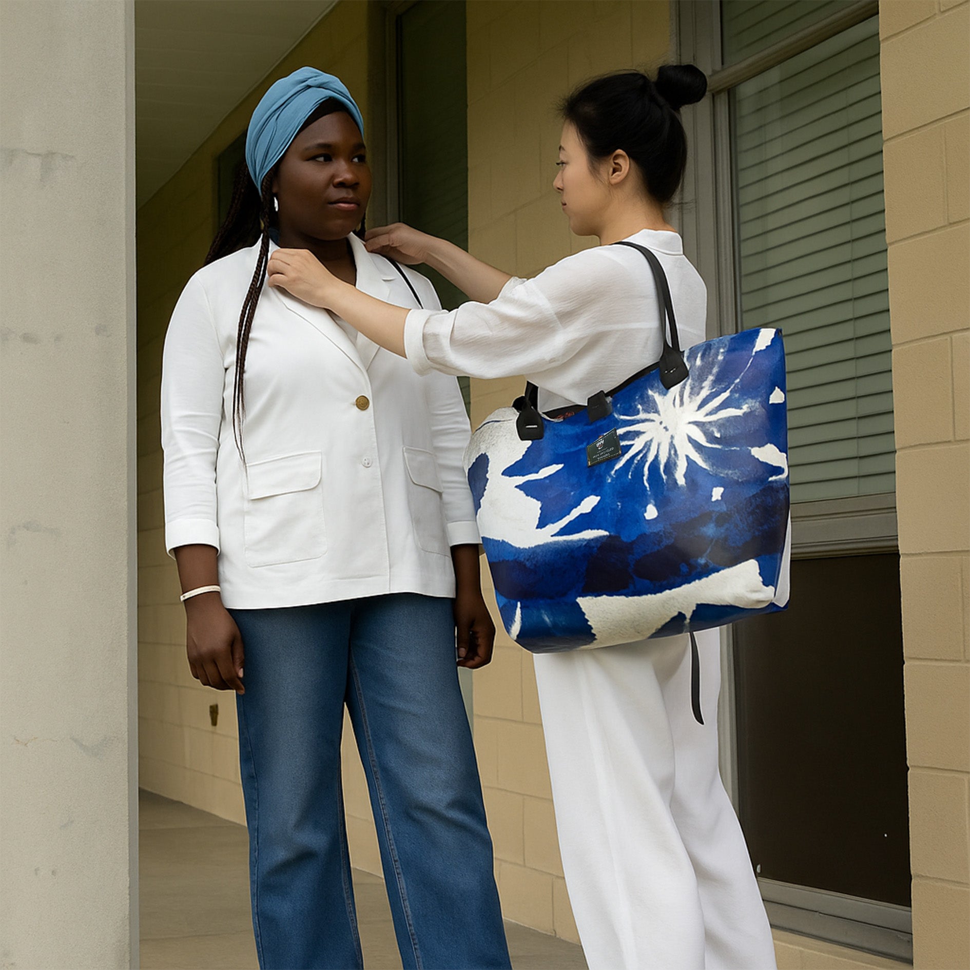 Two women standing outside a building, one adjusting the collar of the other's white coat and holding bright navy blue shopper.